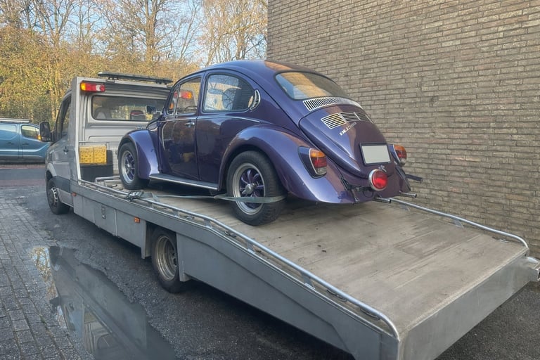 Blue Volkswagen Beetle being transported on a flatbed tow truck parked against a brick wall